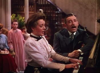Movie still from “Meet Me in St. Louis” (1944), directed by Vincente Minnelli – An older man and a woman playing the piano in front of other people; Medium shot, High angle