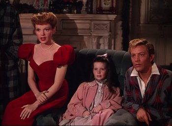 Movie still from “Meet Me in St. Louis” (1944), directed by Vincente Minnelli – A woman in a red dress sitting on a couch next to a man and a little girl; Medium shot, Low angle