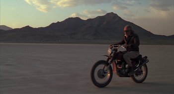 Movie still from “Melvin and Howard” (1980), directed by Jonathan Demme – A man riding a motorcycle in the middle of the desert; Wide shot, Low angle