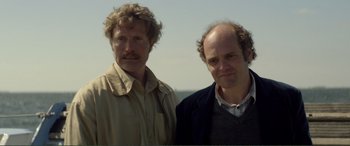 Movie still from “Men & Chicken” (2015), directed by Anders Thomas Jensen – Two men standing next to each other in front of a body of water; Close Up shot, Low angle