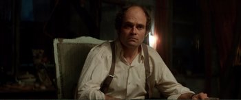 Movie still from “Men & Chicken” (2015), directed by Anders Thomas Jensen – A man sitting in a chair wearing a white shirt and brown suspenders; Medium shot, Over the shoulder angle