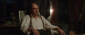 Movie still from “Men & Chicken” (2015), directed by Anders Thomas Jensen – A man sitting in a chair wearing suspenders; Medium shot, High angle