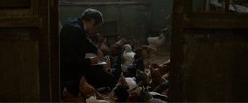 Movie still from “Men & Chicken” (2015), directed by Anders Thomas Jensen – A man sitting on the ground writing on a piece of paper in front of a flock of chickens; Medium shot, High angle