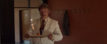 Movie still from “Men & Chicken” (2015), directed by Anders Thomas Jensen – A man in a white suit holding a plate with a cake on top of it; Medium shot, Over the shoulder angle