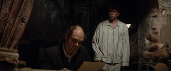 Movie still from “Men & Chicken” (2015), directed by Anders Thomas Jensen – Two men are sitting at a table and one of them is reading a paper; Medium shot, Over the shoulder angle