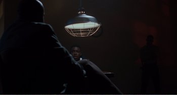 Movie still from “Menace II Society” (1993), directed by Albert Hughes – A man sitting at a table in a dark room; Medium shot, Over the shoulder angle