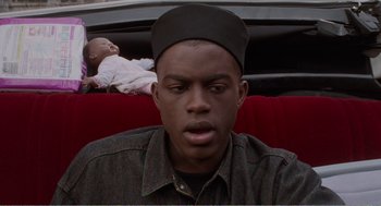 Movie still from “Menace II Society” (1993), directed by Albert Hughes – A young man with a hat on and a baby in the background; Close Up shot, High angle