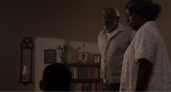 Movie still from “Menace II Society” (1993), directed by Albert Hughes – An older man standing in a living room next to two women; Medium shot, Over the shoulder angle
