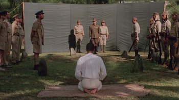 Movie still from “Merry Christmas Mr. Lawrence” (1983), directed by Nagisa Ôshima – A group of men standing around a man in white; Wide shot, High angle