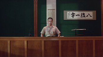 Movie still from “Merry Christmas Mr. Lawrence” (1983), directed by Nagisa Ôshima – A man standing behind a counter in front of a painting; Wide shot, Low angle
