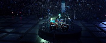 Movie still from “Metallica Through the Never” (2013), directed by Nimród Antal – A man playing drums on a stage in front of an audience; Wide shot, Overhead angle