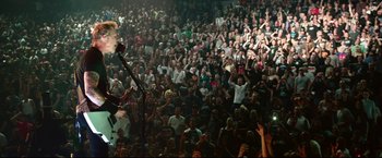 Movie still from “Metallica Through the Never” (2013), directed by Nimród Antal – A crowd of people in a large room with a man on a stage; Wide shot, High angle