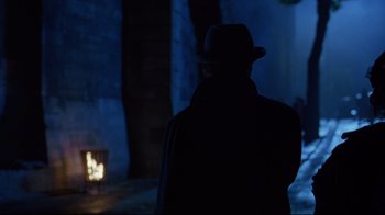 Movie still from “Michael Collins” (1996), directed by Neil Jordan – A man wearing a hat standing in front of a fireplace; Medium shot, Over the shoulder angle