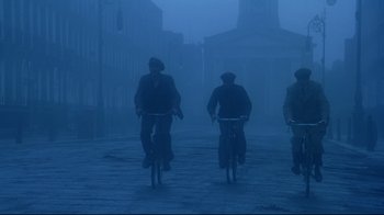 Movie still from “Michael Collins” (1996), directed by Neil Jordan – A group of men riding bikes down a foggy street; Wide shot, Low angle