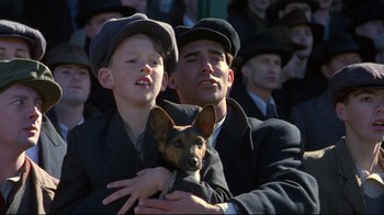 Movie still from “Michael Collins” (1996), directed by Neil Jordan – A man and a boy holding a dog in front of a crowd of onlookers; Medium shot, Over the shoulder angle