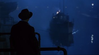 Movie still from “Michael Collins” (1996), directed by Neil Jordan – A man wearing a hat looking at a boat in the water; Wide shot, Over the shoulder angle
