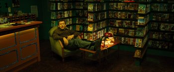 Movie still from “Micmacs” (2009), directed by Jean-Pierre Jeunet – A man sitting on a chair in a room full of movies; Wide shot, High angle
