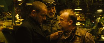 Movie still from “Micmacs” (2009), directed by Jean-Pierre Jeunet – A group of men standing next to each other in a room; Close Up shot, Over the shoulder angle
