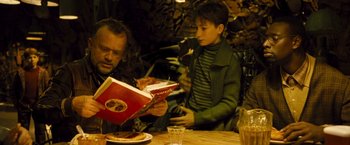 Movie still from “Micmacs” (2009), directed by Jean-Pierre Jeunet – A man and a boy sitting at a table with a book; Medium shot, High angle