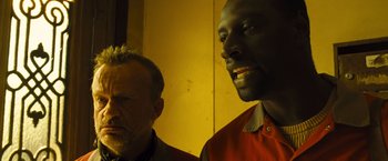Movie still from “Micmacs” (2009), directed by Jean-Pierre Jeunet – Two men standing next to each other in front of a yellow wall; Close Up shot, Low angle