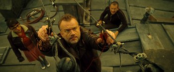 Movie still from “Micmacs” (2009), directed by Jean-Pierre Jeunet – A man holding a bow and arrow while sitting on a bench; Medium shot, Overhead angle