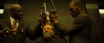 Movie still from “Micmacs” (2009), directed by Jean-Pierre Jeunet – A man is being held up by two men holding guns; Medium shot, High angle