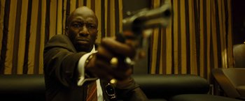 Movie still from “Micmacs” (2009), directed by Jean-Pierre Jeunet – A man in a suit and tie holding a gun in his hand; Close Up shot, Low angle