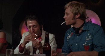 Movie still from “Midnight Cowboy” (1969), directed by John Schlesinger – A couple of men sitting at a table with a bottle of beer and a cigarette; Medium shot, Low angle