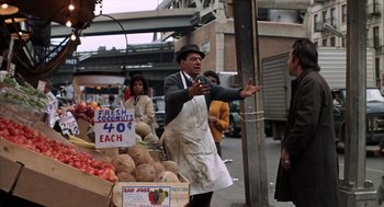 Movie still from “Midnight Cowboy” (1969), directed by John Schlesinger – A man in an apron and a hat standing in front of a fruit stand; Medium shot, Over the shoulder angle