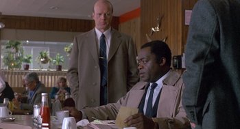 Movie still from “Midnight Run” (1988), directed by Martin Brest – Two men are sitting at a table and one of the men is holding papers; Medium shot, Over the shoulder angle