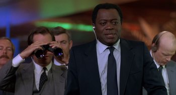 Movie still from “Midnight Run” (1988), directed by Martin Brest – A man in a suit and tie looking through binoculars; Medium shot, Over the shoulder angle