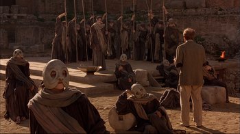 Movie still from “Mighty Aphrodite” (1995), directed by Woody Allen – A group of people in masks are sitting on the ground; Wide shot, High angle