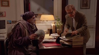 Movie still from “Mighty Aphrodite” (1995), directed by Woody Allen – Two people sitting at a table with a mask on; Medium shot, Over the shoulder angle