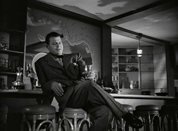Movie still from “Mildred Pierce” (1945), directed by Michael Curtiz – A black and white photo of a man sitting in a chair with a drink; Medium shot, Low angle