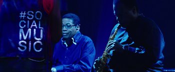 Movie still from “Miles Ahead” (2015), directed by Don Cheadle – A man sitting next to a man playing a saxophone; Medium shot, Low angle