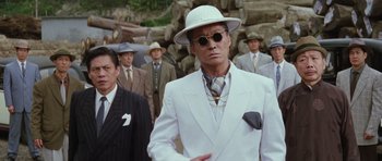 Movie still from “Miracles: The Canton Godfather” (1989), directed by Jackie Chan – A man in a white suit and hat standing in front of a group of men; Medium shot, Low angle