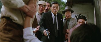 Movie still from “Miracles: The Canton Godfather” (1989), directed by Jackie Chan – A group of men standing next to each other holding a gun; Medium shot, Over the shoulder angle