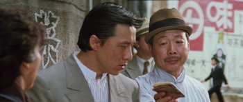 Movie still from “Miracles: The Canton Godfather” (1989), directed by Jackie Chan – A man wearing a suit and hat looking at a sandwich; Close Up shot, Low angle