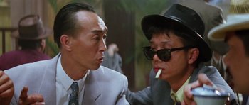 Movie still from “Miracles: The Canton Godfather” (1989), directed by Jackie Chan – A man in a suit and a man in a hat smoking a cigarette; Close Up shot, Low angle
