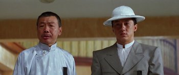 Movie still from “Miracles: The Canton Godfather” (1989), directed by Jackie Chan – A man in a suit and a hat standing next to another man in a suit; Close Up shot, Low angle