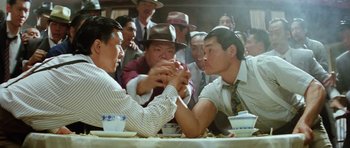 Movie still from “Miracles: The Canton Godfather” (1989), directed by Jackie Chan – Two men arm wrestling at a table with other people; Medium shot, High angle