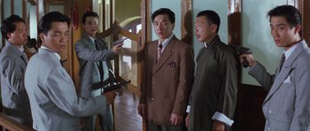 Movie still from “Miracles: The Canton Godfather” (1989), directed by Jackie Chan – A group of men standing next to each other in a room; Medium shot, Over the shoulder angle