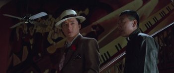 Movie still from “Miracles: The Canton Godfather” (1989), directed by Jackie Chan – A man in a suit and hat standing in front of a staircase; Close Up shot, Low angle