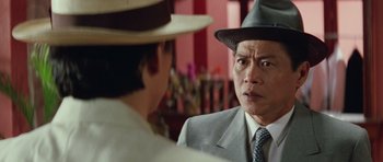 Movie still from “Miracles: The Canton Godfather” (1989), directed by Jackie Chan – A man wearing a suit and hat talking to another man; Close Up shot, Over the shoulder angle