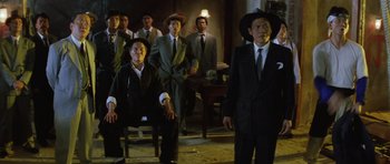 Movie still from “Miracles: The Canton Godfather” (1989), directed by Jackie Chan – A group of men in suits and hats standing in a room; Wide shot, Low angle