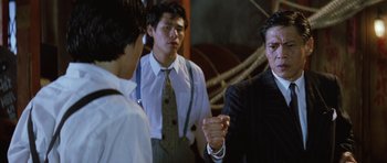 Movie still from “Miracles: The Canton Godfather” (1989), directed by Jackie Chan – A group of men standing next to each other in a room; Medium shot, Over the shoulder angle