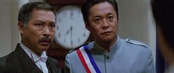 Movie still from “Miracles: The Canton Godfather” (1989), directed by Jackie Chan – A man in a uniform standing next to another man in front of a clock; Close Up shot, Low angle