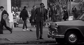 Movie still from “Mirage” (1965), directed by Edward Dmytryk – A black and white photo of a man standing next to an old car; Wide shot, Over the shoulder angle
