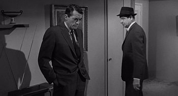 Movie still from “Mirage” (1965), directed by Edward Dmytryk – A man in a suit and a man in a hat standing next to each other in a room; Medium shot, Over the shoulder angle