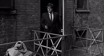 Movie still from “Mirage” (1965), directed by Edward Dmytryk – A man in a suit and hat standing in a doorway; Medium shot, Over the shoulder angle